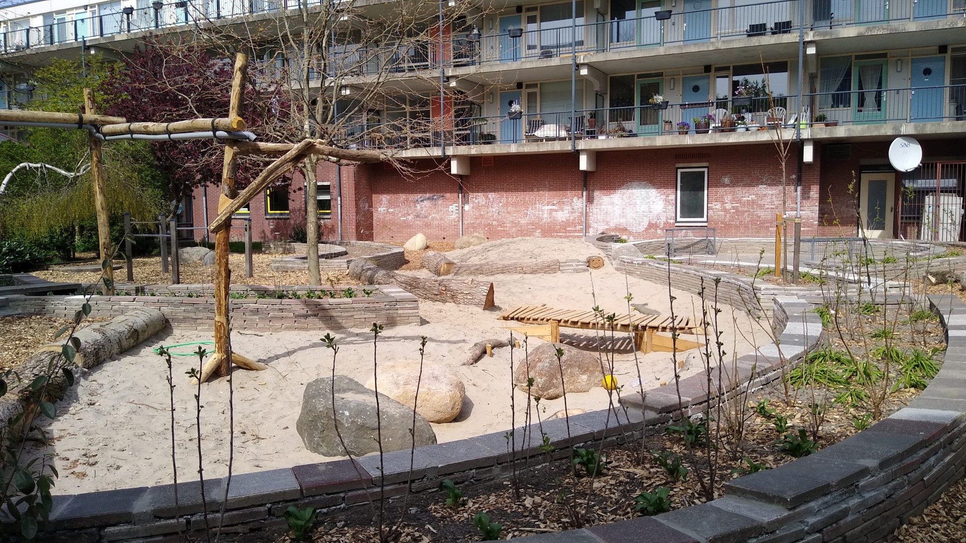 Zandspeelplek met heuvels, boomstammen en gemetselde randen tussen flats